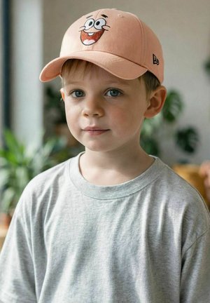 Junge mit blauen Augen, der ein hellgraues Hemd und eine pfirsichfarbene Mütze mit einem lächelnden Cartoon-Gesicht trägt, drinnen mit verschwommenen Pflanzen im Hintergrund.