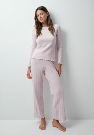 Jeune femme debout pieds nus contre un mur blanc, portant un haut à manches longues rose clair et un pantalon large assorti avec un motif à carreaux subtil.