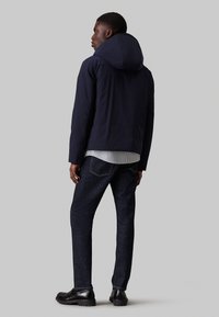 Giacca con cappuccio blu navy dalla texture liscia, abbinata a una maglietta a righe sotto e jeans scuri in denim. Le scarpe nere completano il look.