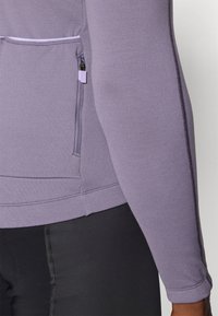 Top de manga larga lavanda con un acabado texturizado, que presenta un bolsillo lateral con cremallera y detalles cosidos, combinado con leggings negros.