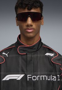 Black racing suit with red accents and white Formula 1 logo, featuring a high collar and protective stitching. Dark sunglasses complete the look.