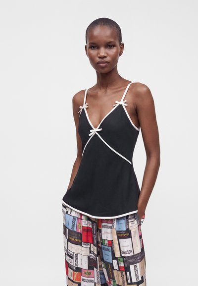 Femme aux cheveux courts portant un caraco noir avec des bordures blanches et des nœuds, associé à un pantalon large coloré avec des motifs d'étiquettes.
