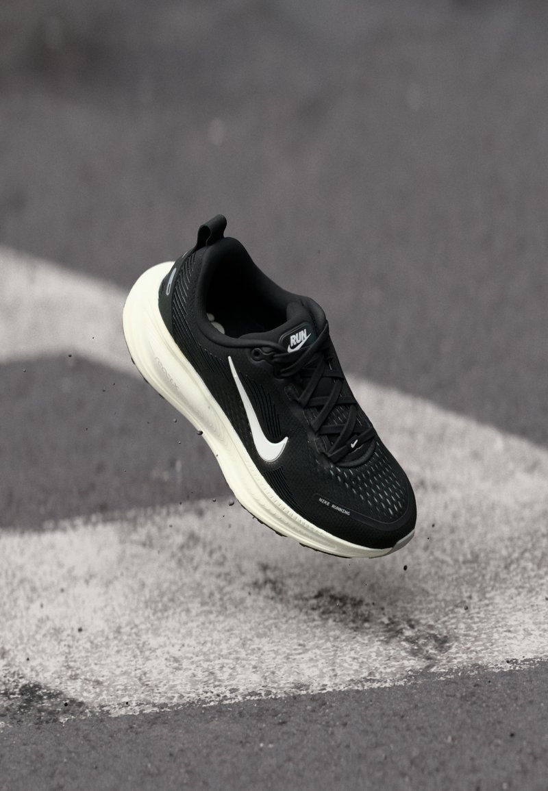 Zapatilla de running negra con una superficie texturizada, swoosh blanco y suela color crema, que cuenta con un tirador y cordones para un ajuste seguro.