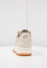 Beige Nike Air-sneaker med texturerat sidomönster och gummisole, sedd bakifrån på vit yta med helt vit bakgrund.