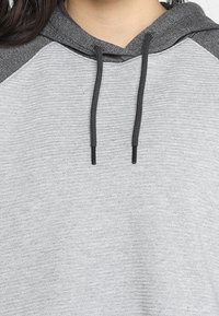 Sweat-shirt à capuche gris avec un corps texturé, des manches raglan gris foncé et des cordons. Présente une coupe décontractée et un design minimaliste.