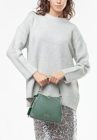 Borsa a mano in pelle verde con design strutturato, manico corto e accenti in hardware argentato lucido. Indossati un maglione grigio a trama e una gonna con paillettes.