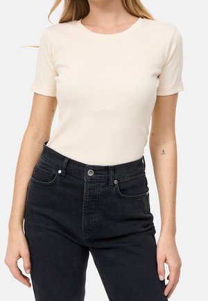Vrouw draagt een aansluitend, roomkleurig, geribd kortärmelig shirt, gestopt in een high-waisted zwarte spijkerbroek, tegen een witte achtergrond.