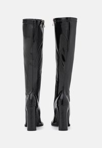 Bottes noires montant jusqu'au genou en cuir verni brillant, avec un talon bloc et une fermeture éclair latérale pour faciliter l'enfilage. Texture lisse et design élégant.