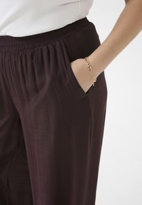 Wide-leg, purple pants made from a textured fabric with an elastic waistband and side pockets. A gold chain bracelet is visible on the wrist.