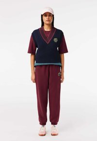 Pull en maille sans manche bleu marine avec accents rouges, superposé sur une chemise à manches courtes bordeaux ; associé à un pantalon de survêtement bordeaux et des baskets roses.