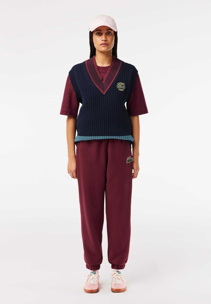 Pull en maille sans manche bleu marine avec accents rouges, superposé sur une chemise à manches courtes bordeaux ; associé à un pantalon de survêtement bordeaux et des baskets roses.