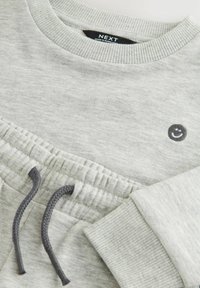 Haut de sweatshirt gris clair avec col et poignets côtelés, orné d'un petit détail brodé de visage souriant. Tissu doux avec une coupe décontractée.