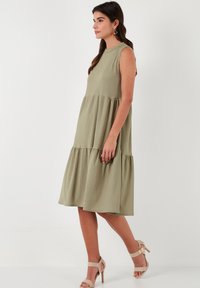 Light green sleeveless tiered dress made of textured fabric, featuring a round neckline and a flowing A-line shape, worn with beige sandals.