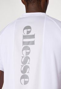 Weißes kurzärmliges Hemd mit vertikalem grauen Logo-Druck auf dem Rücken und einem strukturierten Mesh-Panel. Glatte Stoffqualität mit rundem Ausschnitt.
