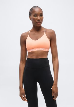 Kvinde iført en ferskenfarvet Nike sports-bh og sorte, højtaljede leggings, stående mod en ensfarvet lyse baggrund.