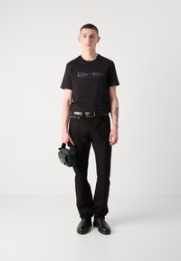 Sort t-shirt med "Calvin Klein" logo, sorte jeans, sort bælte og sorte sko. Holder en lille, struktureret sort taske.