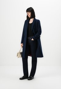 Marineblauer Wollmantel mit einem einzigen Knopf, kombiniert mit dunklen Jeans, schwarzem Oberteil und schwarzen Slippern. Hält eine kleine, hellfarbige Handtasche.
