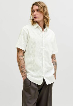 Junger Mann mit langen blonden Haaren trägt ein weißes kurzärmeliges Hemd mit Knöpfen und lockere dunkle Hose, steht mit den Händen in den Taschen und schaut seitlich.