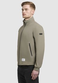 Világos olíva zöld kabát magas gallérral, oldalsó zipzáras zsebbel és rugalmas mandzsettákkal. Bal vállán logó patch található, minimalistára tervezve.