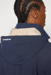 Giacca navy con cappuccio beige chiaro e cuciture, con un logo sul colletto posteriore. Tessuto liscio e non testurizzato con un taglio moderno.