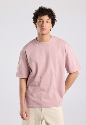 Jong persoon met krullend haar, gekleed in een losse roze t-shirt en beige broek, staand tegen een eenvoudige lichte achtergrond.