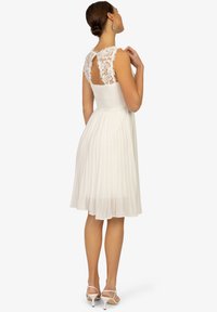 Weißes Kleid mit tailliertem, gerafftem Oberteil und plissiertem Rock. Der Rücken verfügt über Spitzenverzierungen mit einer Schlüsselöffnung und einem fließenden Design.