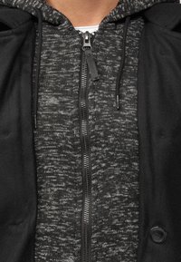 Grå og svart zip-up hettegenser med teksturert stoff, svarte detaljer og snor i hetten. Har full glidelås og kontrasterende sømmer.