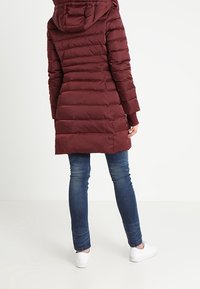 Kvinna iförd en lång burgundy vadderad jacka med huva, blå jeans och vita sneakers, stående framför en enfärgad vit bakgrund.