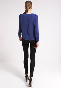 Ruskeatukkainen nainen poninhännällä, pukeutunut siniseen pitkähihalaiseen puseroon, mustiin kapealahkeisiin farkkuihin ja mustiin korkokenkiin, seisoo valkoista taustaa vasten.