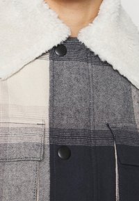 Kurtka-koszula w kratę w kolorach granatowym, szarym i kremowym, z białym kołnierzem z sztucznego baranka i kieszenią na piersi.