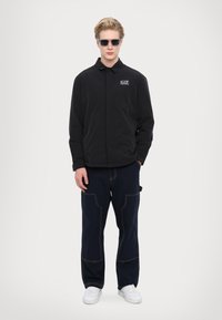 Svart lättviktsjacka med krage, prydd med "EA7"-logotyp. Kombinerad med marinblå byxor med kontrasterande röda stygn och vita sneakers.