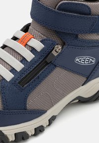 Keen avalynė, turinti tamsiai mėlynos odos ir pilko tekstūruoto audinio viršutinę dalį, baltus raištelius, oranžinį akcentą ir užtrauktuko detalę šone.