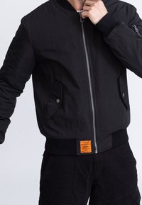 Homme portant une veste bomber noire avec fermeture éclair et poches pressionnées, ajustant le col d'une main.