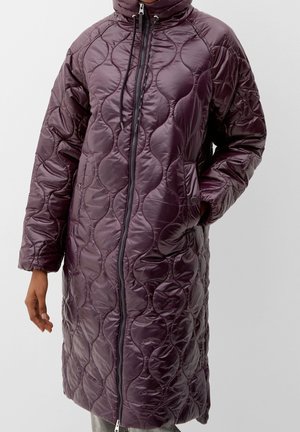 Longue veste matelassée violette avec col montant, fermeture éclair à l'avant, cordon de serrage, poches latérales, une main dans une poche, portée par une personne sur un fond uni.