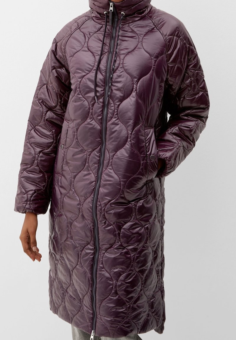 Longue veste matelassée violette avec col montant, fermeture éclair à l'avant, cordon de serrage, poches latérales, une main dans une poche, portée par une personne sur un fond uni.