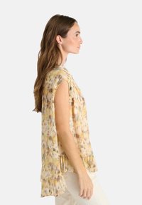 Mujer de perfil con una blusa suelta sin mangas y volantes con un patrón abstracto en amarillo y melocotón, y pantalones de color claro.