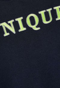 T-shirt en coton navy avec un design brodé portant le mot "UNIQUE" en lettres vert fluo. Texture lisse sans motifs visibles.