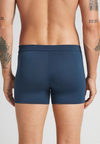 Mörkblå trikåshorts med en mjuk textur, elastisk midja och minimala sömmar. Bärs av en person sett bakifrån, vilket visar tatuerade armar.