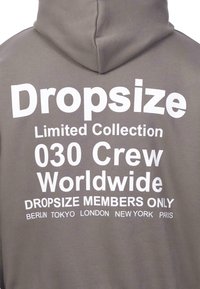 Grauer Hoodie mit fettem weißen Text, der "Dropsize Limited Collection", "030 Crew Worldwide" und Städtenamen wie Berlin, Tokio, London, New York und Paris beinhaltet.