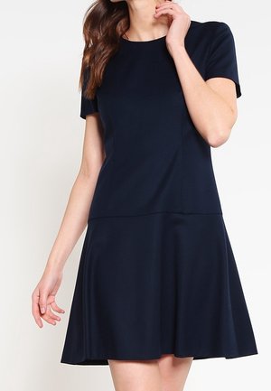 Mujer vestida con un vestido de manga corta, azul marino, hasta la rodilla, con un corpiño ajustado y falda acampanada, posando frente a un fondo liso.