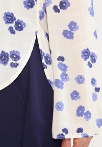 Blouse légère en crème à motifs floraux bleus. Dotée de manches évasées et d'une fente sur le côté. Associée à un tissu marine en dessous.