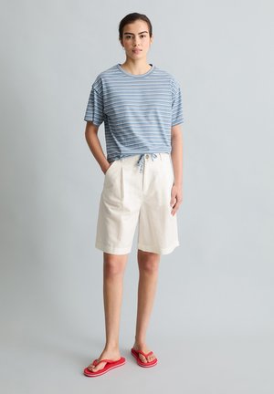 Młoda kobieta stojąca z rękami w kieszeniach, ubrana w niebieską koszulkę w paski, białe szorty do kolan i czerwone klapki, na jednolitym tle.