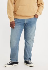Light blue denim jeans with a relaxed fit, featuring a straight leg design, classic five-pocket styling, worn with a beige hoodie and black sneakers.