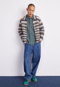 Flerfärgad randig cardigan i nyanser av brunt och grått, bärs över en grön rutad skjorta, ihop med blå lösa jeans och gröna sneakers.