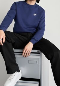 Námořnický modrý svetřík s kulatým výstřihem má bílé logo Nike, v kombinaci s černými kalhotami a bílými teniskami, situovaný na neutrálním pozadí.