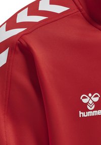Giacca sportiva rossa realizzata in tessuto liscio, con motivi a chevron bianchi lungo le spalle e un logo bianco sul petto.