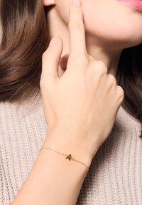 Złota bransoletka z zawieszką w kształcie litery "A", delikatny łańcuszek, noszona na nadgarstku. Tło stanowi teksturowana beżowa dzianina.