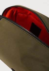 Τσάντα με φερμουάρ σε ελαιί πράσινο χρώμα και μαύρο φερμουάρ, με φωτεινή κόκκινη επένδυση στο εσωτερικό και μια μικρή τσέπη για οργάνωση.