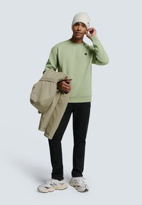 Lichtgroene sweatshirt met ronde hals en embleem, beige buitenjasje over de arm, zwarte broek en witte sneakers met grijze accenten.