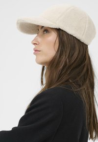 Cream fleece cap with a rounded visor, featuring a textured surface and seam detailing. Model wears a black coat, hair partially visible.